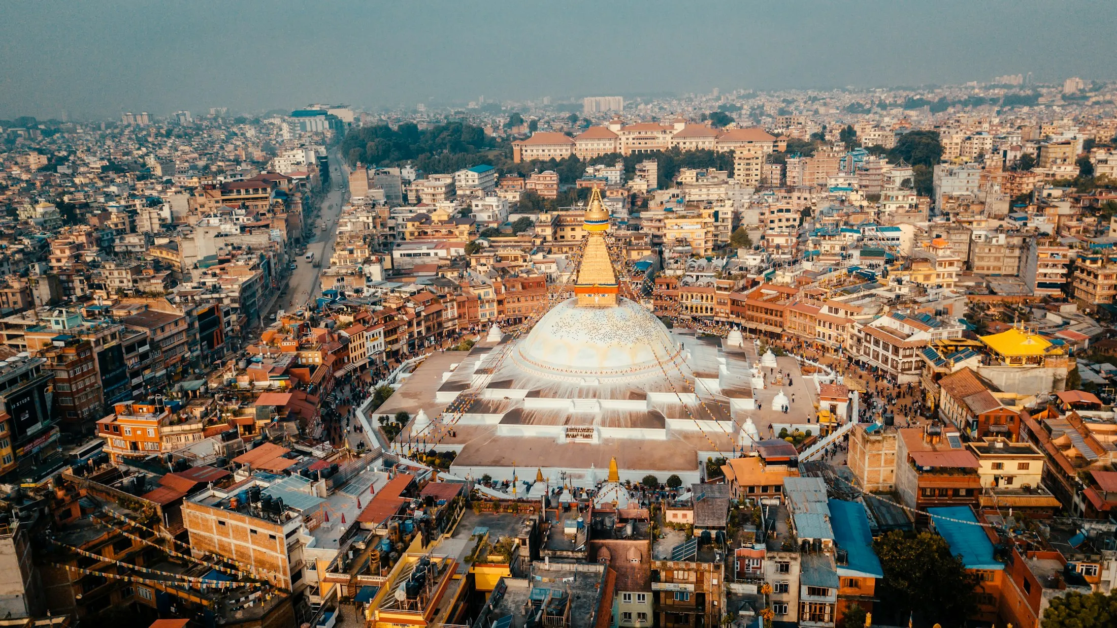 Aerial view of Boudhanath stupa in golden afternoon light, surrounded by the rooftops of Boudha